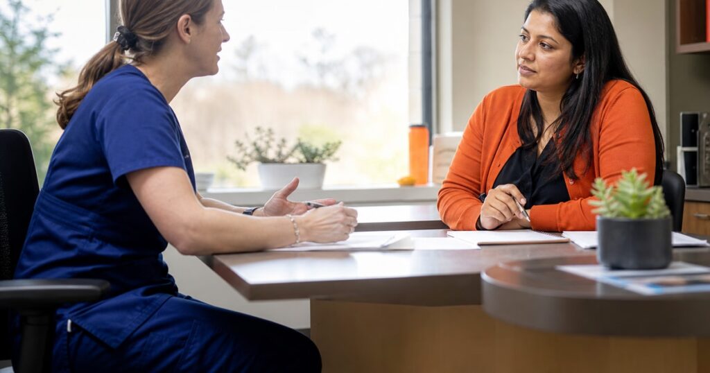 A nurse practitioner and a hiring manager sit across a desk in a clinic office, mid-conversation, papers and notes between them as afternoon light fills the room from behind. · i4 Search Group